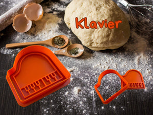 Orange Klavier Keks-Ausstecher auf Backunterlage mit Teig und Zutaten. Ideal für kreative Backprojekte.