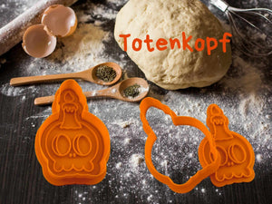Orange Totenkopf-Ausstecher für Halloween-Kekse neben Teig und Backzubehör auf dunklem Untergrund