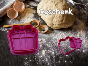 Gift-shaped cookie cutter in pink on a baking surface with dough and kitchen utensils.